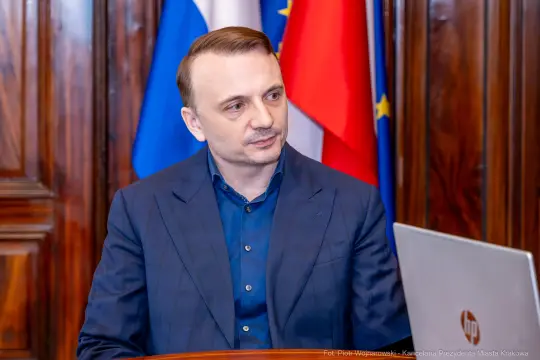 Mężczyzna w granatowej marynarce siedzi przy biurku, w tle flagi.