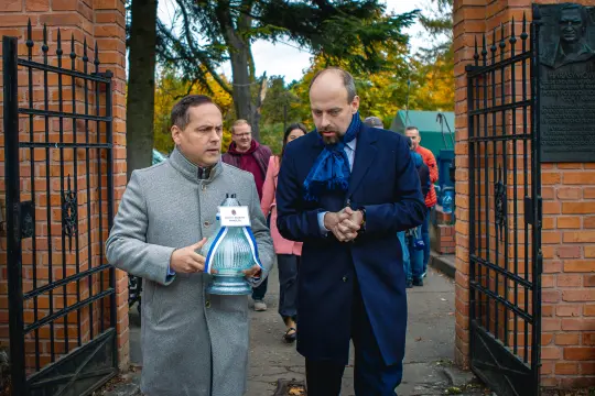 Dwaj mężczyźni niosą lampę w parku, w tle widoczni inni ludzie i brama.