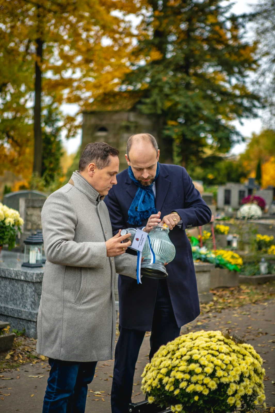 Dwóch mężczyzn na cmentarzu, jeden trzyma lampion, drugi robi zdjęcie. W tle kwiaty i nagrobki.  Autor: Tom Rollauer