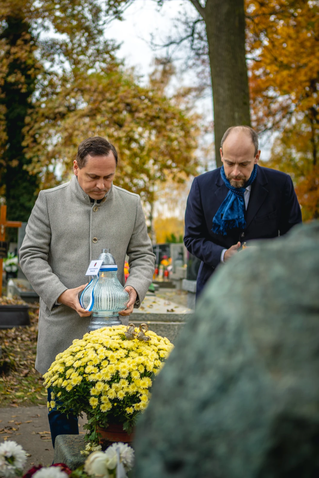 Dwaj mężczyźni z zniczami stoją przy grobie, w tle jesienne liście i kwiaty.  Autor: Tom Rollauer