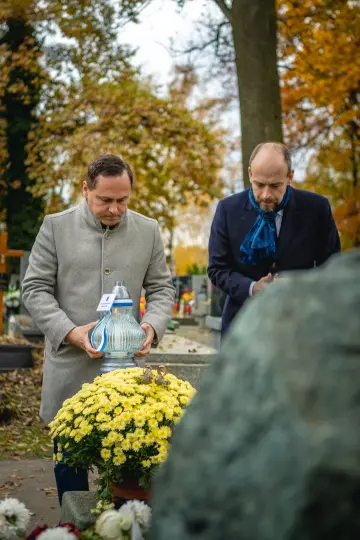 Dwaj mężczyźni z zniczami stoją przy grobie, w tle jesienne liście i kwiaty.
