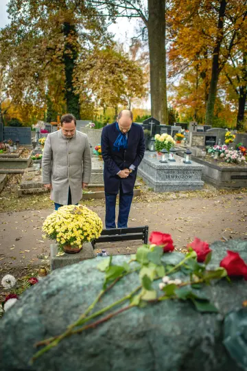Dwóch mężczyzn składa hołd na cmentarzu, obok kwiaty i znicze.