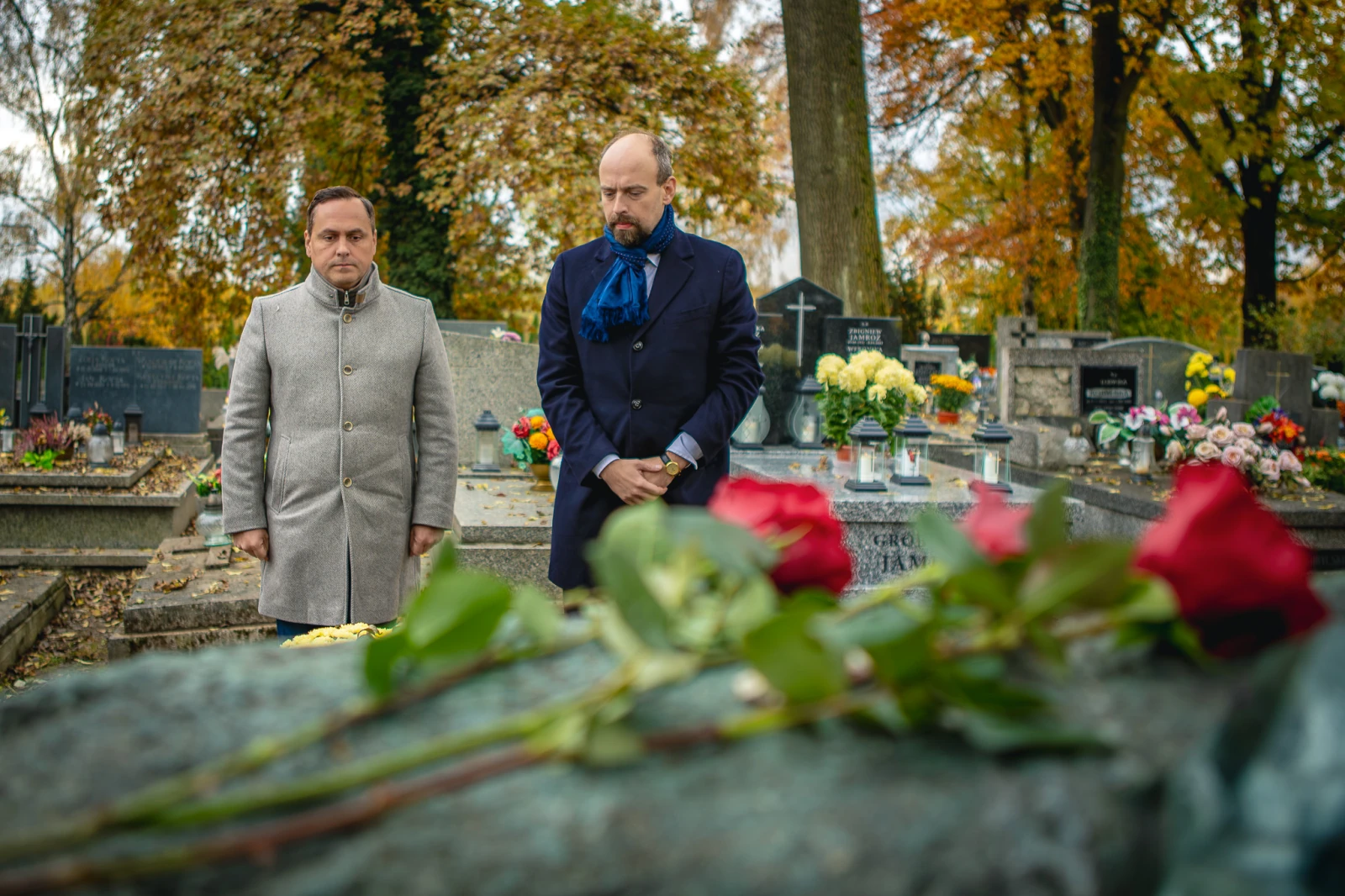 Dwaj mężczyźni w eleganckich ubraniach stoją przy grobie, obok leżą czerwone róże. Jesienna sceneria.  Autor: Tom Rollauer