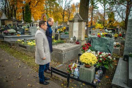 Dwaj mężczyźni stoją przy grobie, otoczonym kwiatami i zniczami, w jesiennej scenerii.