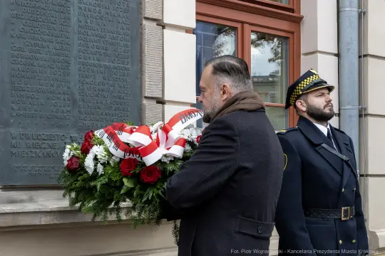 Mężczyzna składa wieniec pod tablicą pamiątkową. Obok stoi funkcjonariusz w mundurze.