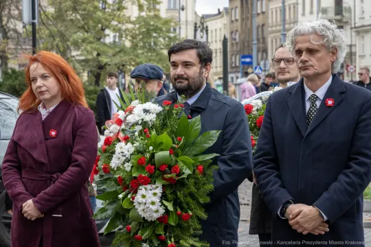 Uczestnicy uroczystości z kwiatami w rękach, w tle ulica i budynki.