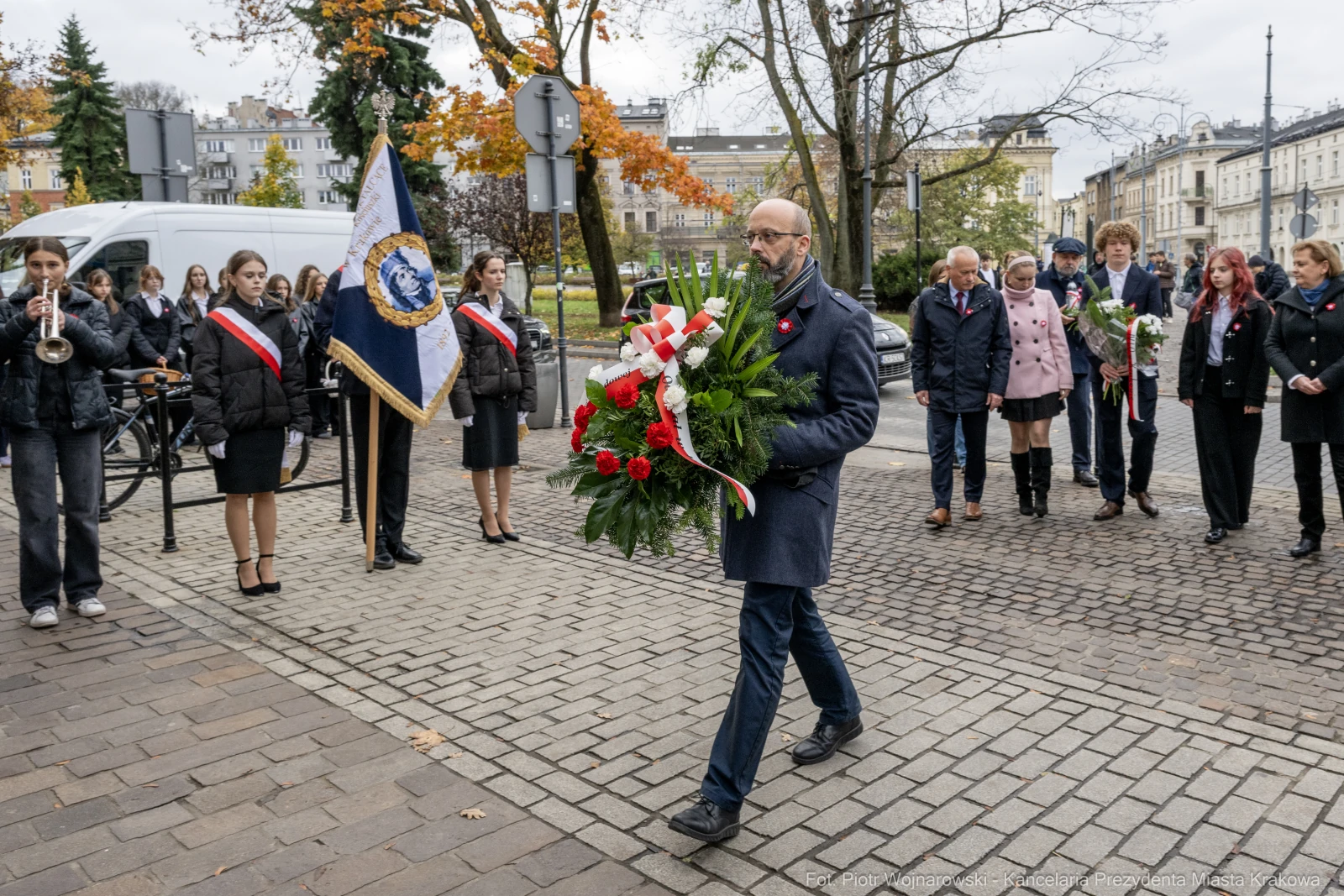 Mężczyzna niosący wieniec kwiatów, towarzyszy mu grupa osób w strojach galowych.  Autor: Piotr Wojnarowski
