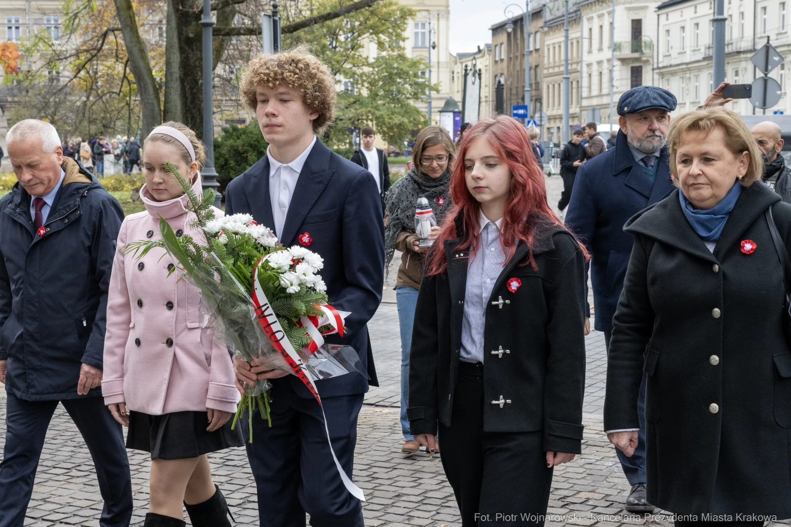 Grupa ludzi w trakcie ceremonii z kwiatami, w strojach formalnych, z symbolami na ubraniach.  Autor: Piotr Wojnarowski