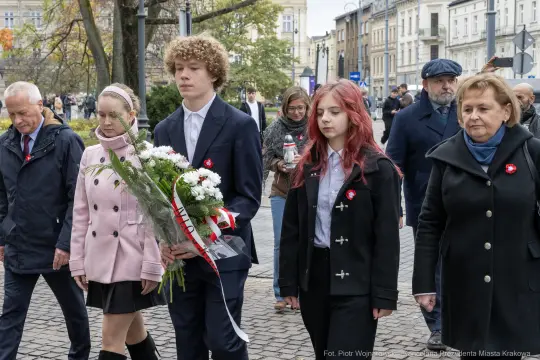 Grupa ludzi w trakcie ceremonii z kwiatami, w strojach formalnych, z symbolami na ubraniach.