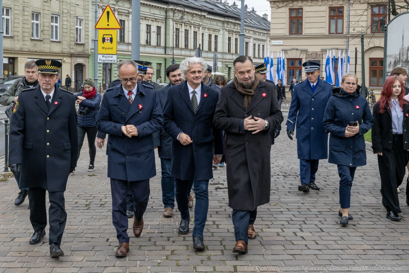 Grupa ludzi w garniturach i mundurach idzie ulicą, niektórzy mają przypięte czerwone kokardy.  Autor: Piotr Wojnarowski