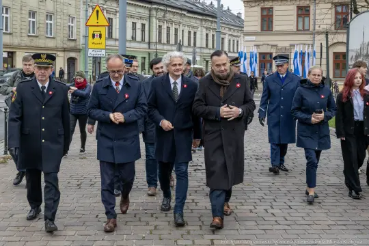 Grupa ludzi w garniturach i mundurach idzie ulicą, niektórzy mają przypięte czerwone kokardy.
