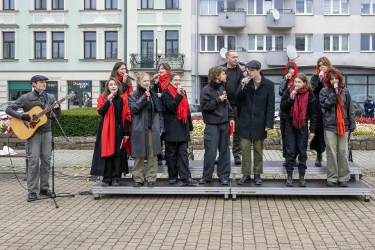 Grupa wykonawców śpiewa na scenie, część z nich trzyma mikrofony, a jeden gra na gitarze.