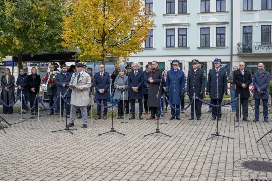 Uroczystość złożenia kwiatów, uczestnicy w eleganckich strojach, jesienna sceneria.