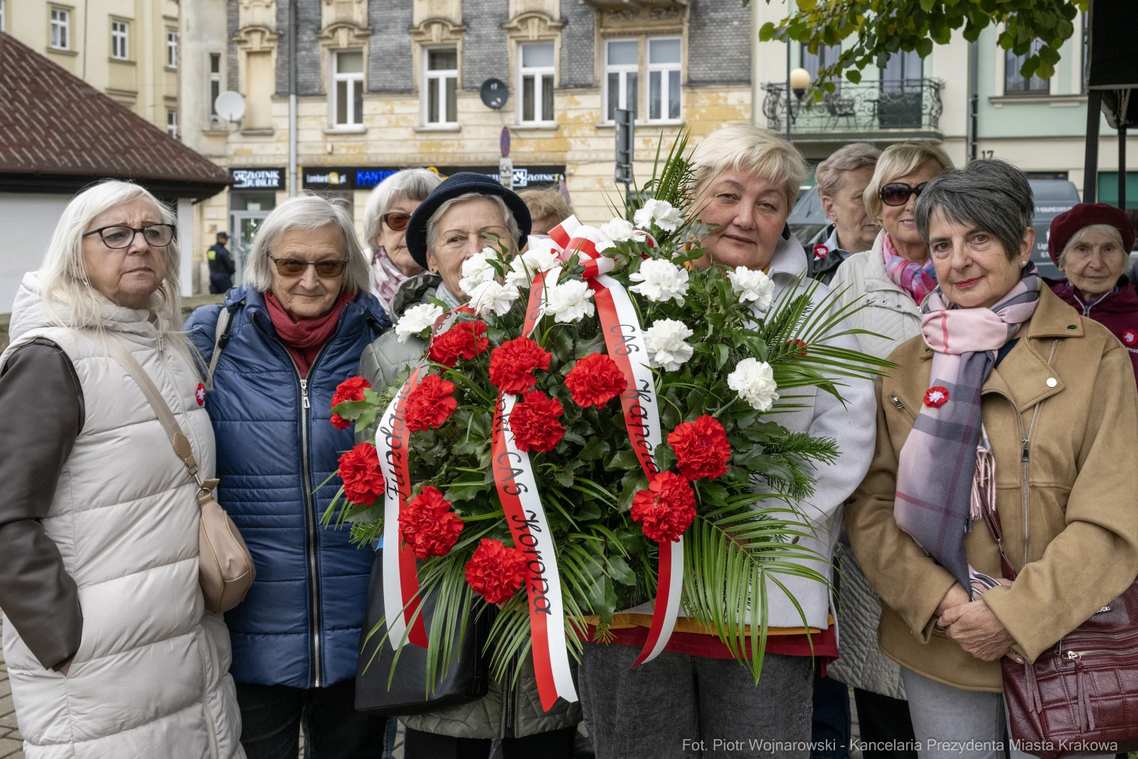 Grupa kobiet z kwiatami i wstążkami, uczestniczących w uroczystości upamiętniającej.  Autor: Piotr Wojnarowski