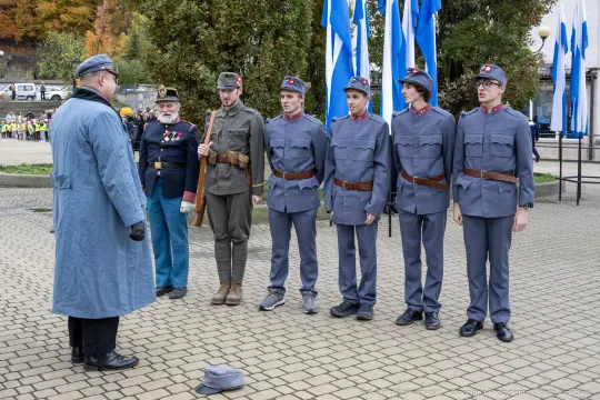 Grupa mężczyzn w mundurach wojskowych podczas ceremonii, z flagami w tle.