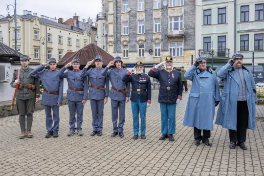 Grupa mężczyzn w mundurach wojskowych, salutujących na tle budynków.