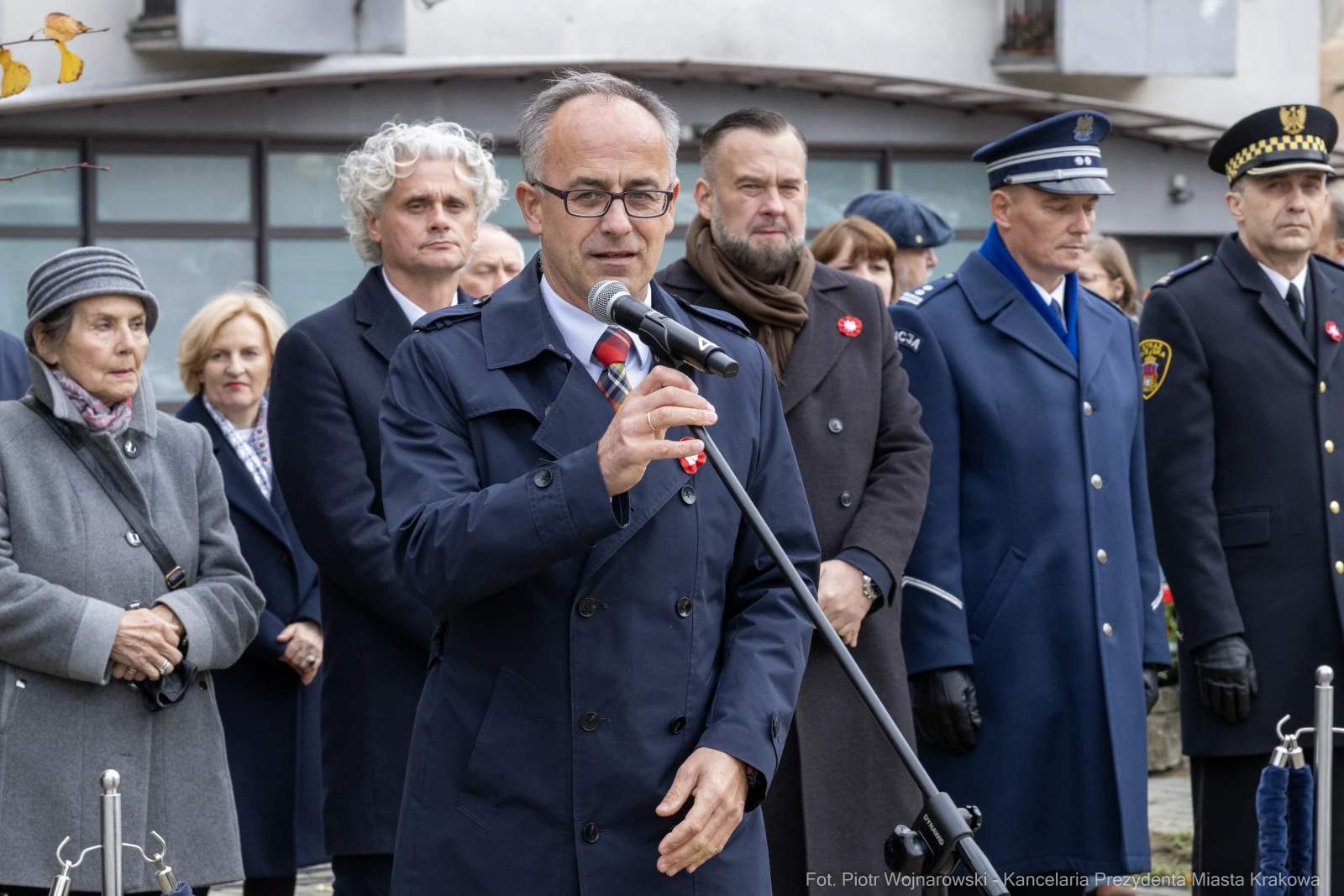 Mężczyzna w okularach przemawia do mikrofonu, w tle grupa osób w mundurach i eleganckich strojach.  Autor: Piotr Wojnarowski