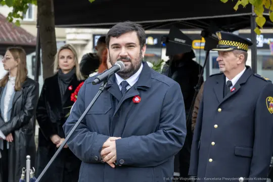 Mężczyzna przemawia do zgromadzenia, w tle osoby w strojach formalnych.