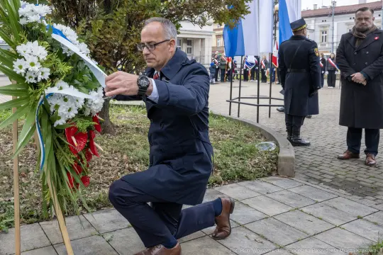 Mężczyzna składa wieniec w hołdzie, w tle flagi i zgromadzenie ludzi.