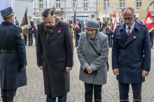 Trzy osoby w eleganckich ubraniach składają hołd, stojąc w milczeniu na uroczystości.