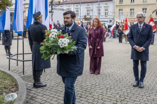 Mężczyzna z bukietem kwiatów na uroczystości z flagami i uczestnikami w tle.
