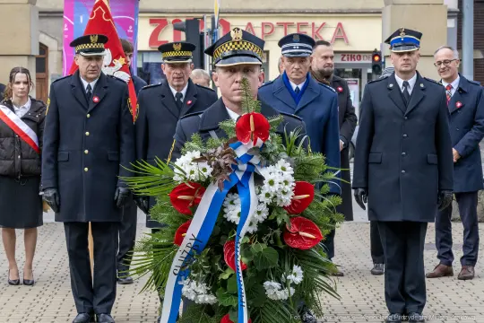 Mężczyzna w mundurze niesie wieniec z kwiatami, obok niego grupa osób w formalnych strojach.