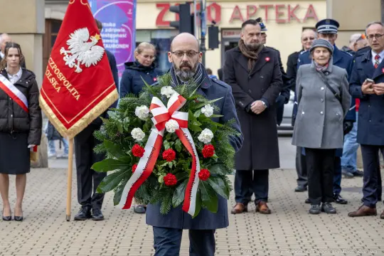 Mężczyzna niesie wieniec z białymi i czerwonymi kwiatami podczas uroczystości.