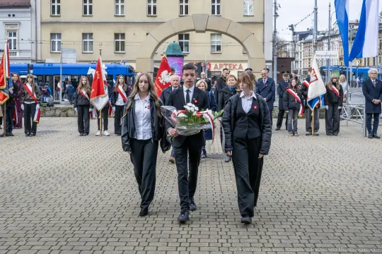 Uczestnicy ceremonii niosący kwiaty w hołdzie, z flagami w tle.