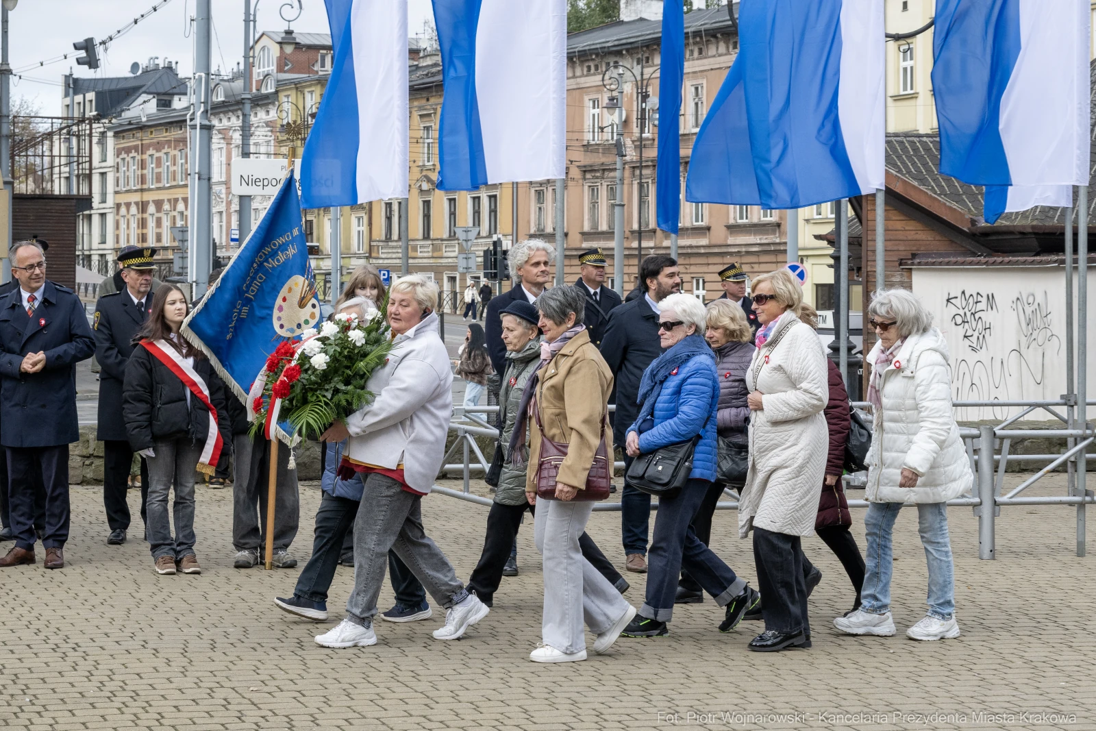 Grupa osób niosąca kwiaty podczas uroczystości, w tle flagi i budynki.  Autor: Piotr Wojnarowski