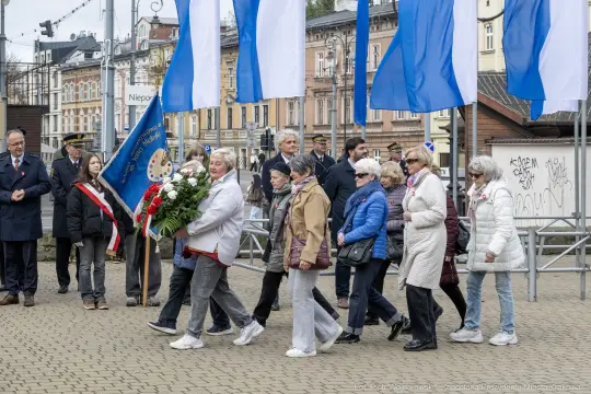 Grupa osób niosąca kwiaty podczas uroczystości, w tle flagi i budynki.