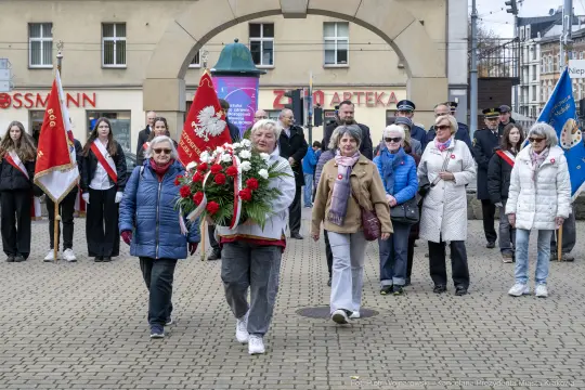 Grupa ludzi niosąca wieniec z flagą Polski podczas uroczystości.
