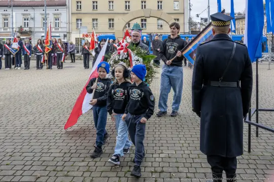 Uczestnicy ceremonii z flagami i wieńcem, dzieci w niebieskich czapkach idą w procesji.