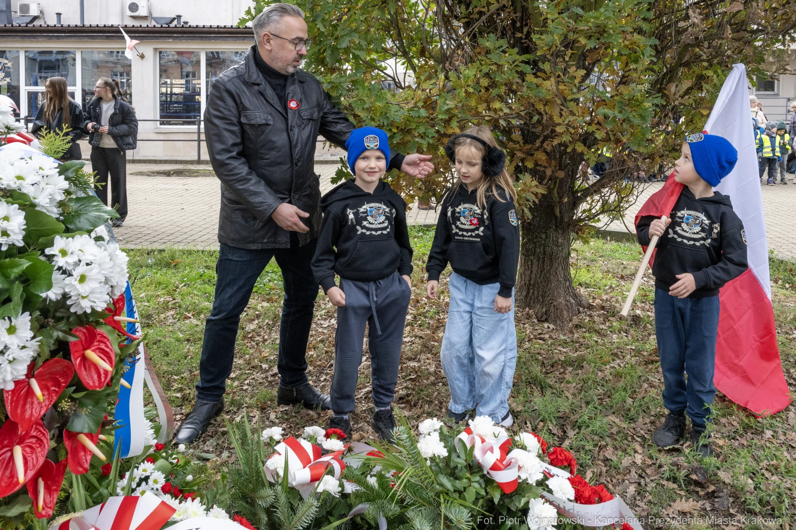 Mężczyzna z dziećmi przy pomniku, składają kwiaty w hołdzie. Dzieci w czapkach i bluzach z logo.  Autor: Piotr Wojnarowski