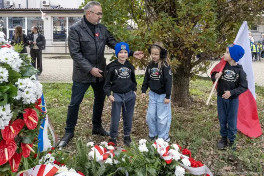 Mężczyzna z dziećmi przy pomniku, składają kwiaty w hołdzie. Dzieci w czapkach i bluzach z logo.