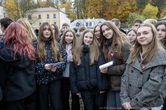 Grupa uczniów w różnych strojach, uśmiechniętych, z kartkami w rękach, w tle kolorowe drzewa.
