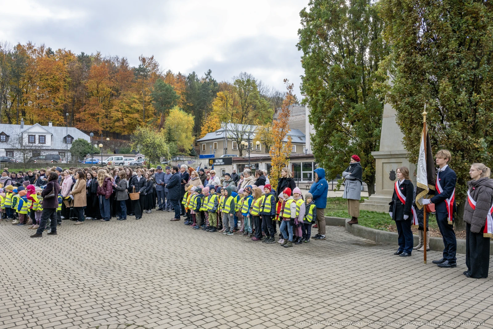 Uczestnicy uroczystości z flagami i dzieci w kamizelkach odblaskowych. Jesienna sceneria.  Autor: Piotr Wojnarowski