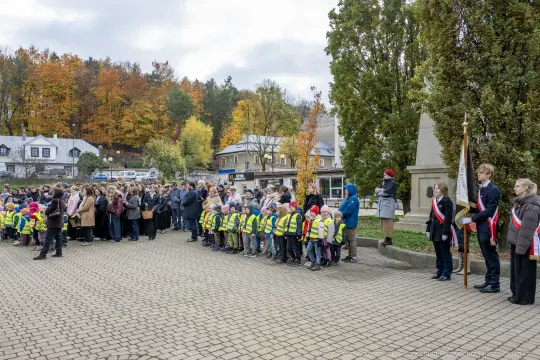 Uczestnicy uroczystości z flagami i dzieci w kamizelkach odblaskowych. Jesienna sceneria.
