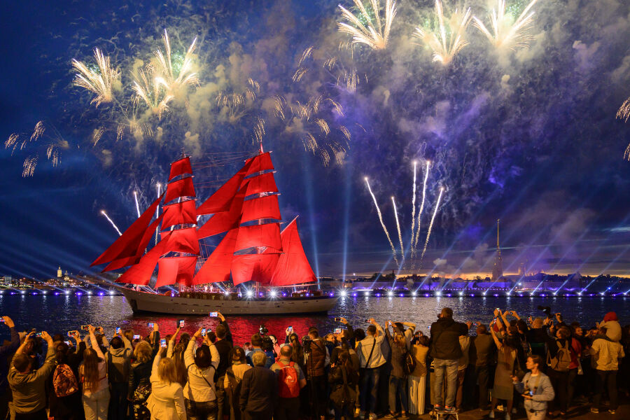 Noc, z portu na morze wypływa duży statek z czerwonymi żaglami. Na statek kierowane są światła reflektorów, na ciemnym niebie sztuczne ognie
