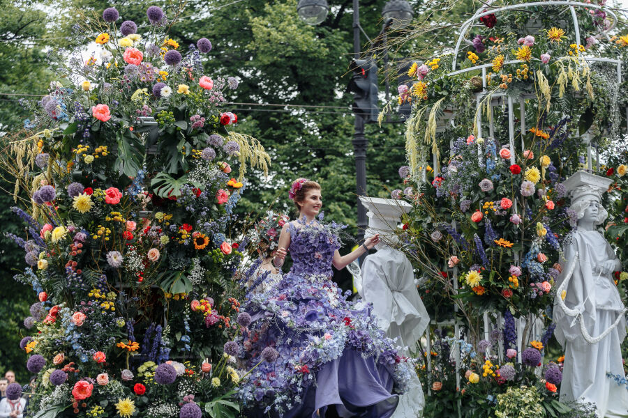 Na tle altany z kolorowych kwiatów stoi kobieta w fioletowej balowej sukni pokrytej kwiatami