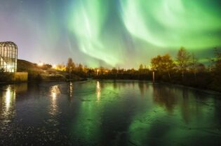 Zorza polarna w Finlandii. Zbiornik wodny i oświetlona konstrukcja ze szkła i stali. Wokół zbiornika wodnego drzewa. Na ciemniejszym  niebie jasnozielone świetliste pasy zorzy polarnej.  