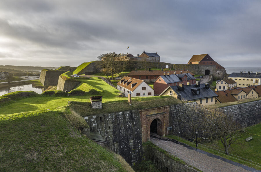 Twierdza Varberg. Na pierwszym planie brama oraz mury i umocnienia ziemne porośnięte zieloną trawą. Dalej po lewej oraz w centrum zabytkowe domy wewnątrz murów.   