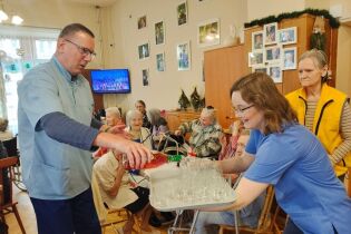 Koniec roku Mężczyzna rozlewa szampana do literatek, kobieta trzyma tacę. Na drugim planie kilkanaście uśmiechniętych osób.