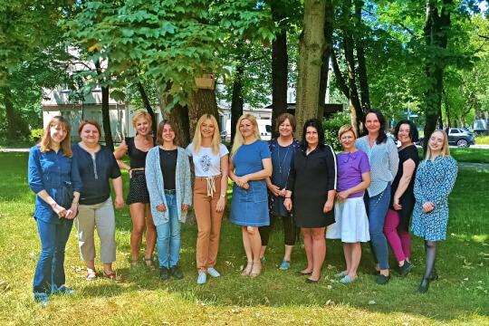 Na fotografii widzimy grupę dwunastu pań stojących na trawie pomiędzy wysokimi zielonymi drzewami. 