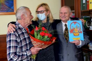 Dzień Kobiet 08.03.2021 trzy uśmiechnięte osoby , kobieta - dyrektor DPS-u obejmująca ramionami dwóch mieszkańców Domu. Jeden z nich trzyma bukiet tulipanów, drugi laurkę okolicznościową.