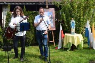 Misyjna Majówka 13.05.2021 Jeden z mężczyzn mówi do mikrofonu, drugi z gitarą w rękach przygotowuje się do prowadzenia śpiewu. Po prawej stronie znajduje się figurka Matki Bożej Fatimskiej.