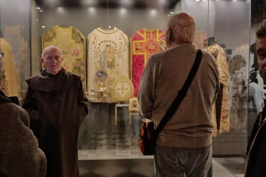 Zdjęcie przedstawia Karmelitę Bosego, w trakcie omawiania historii zakonu. Za jego plecami znajduje się szklana gablota, w której wiszą zabytkowe ornaty liturgiczne.