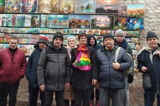 W poszukiwaniu wiosny 21.03.2023 Grupa kobiet i mężczyzn na tle obrazów wiszących na Bramie Floriańskiej. Jedna z kobiet trzyma w ręku marzannę.