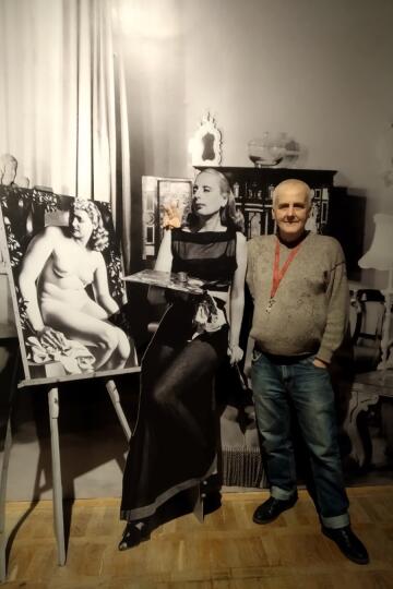 mężczyzna na tle zainscenizowanej pacownii, obok fotografii całopostaciowej artystki
