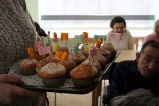 Dzień Kobiet 08.03.2023 Taca z muffinkami trzymana w dłoniach. W tle siedzący kobieta i mężczyzna.