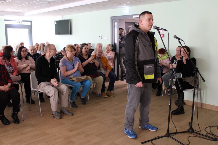 Stojący przy mikrofonie mężczyzna w pomieszczeniu. W tle grupa siedzących  kobiet i mężczyzn.